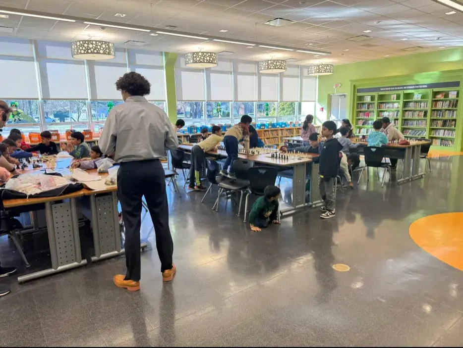 Rows of students playing chess at long tables in a library while an adult walks between the boards.